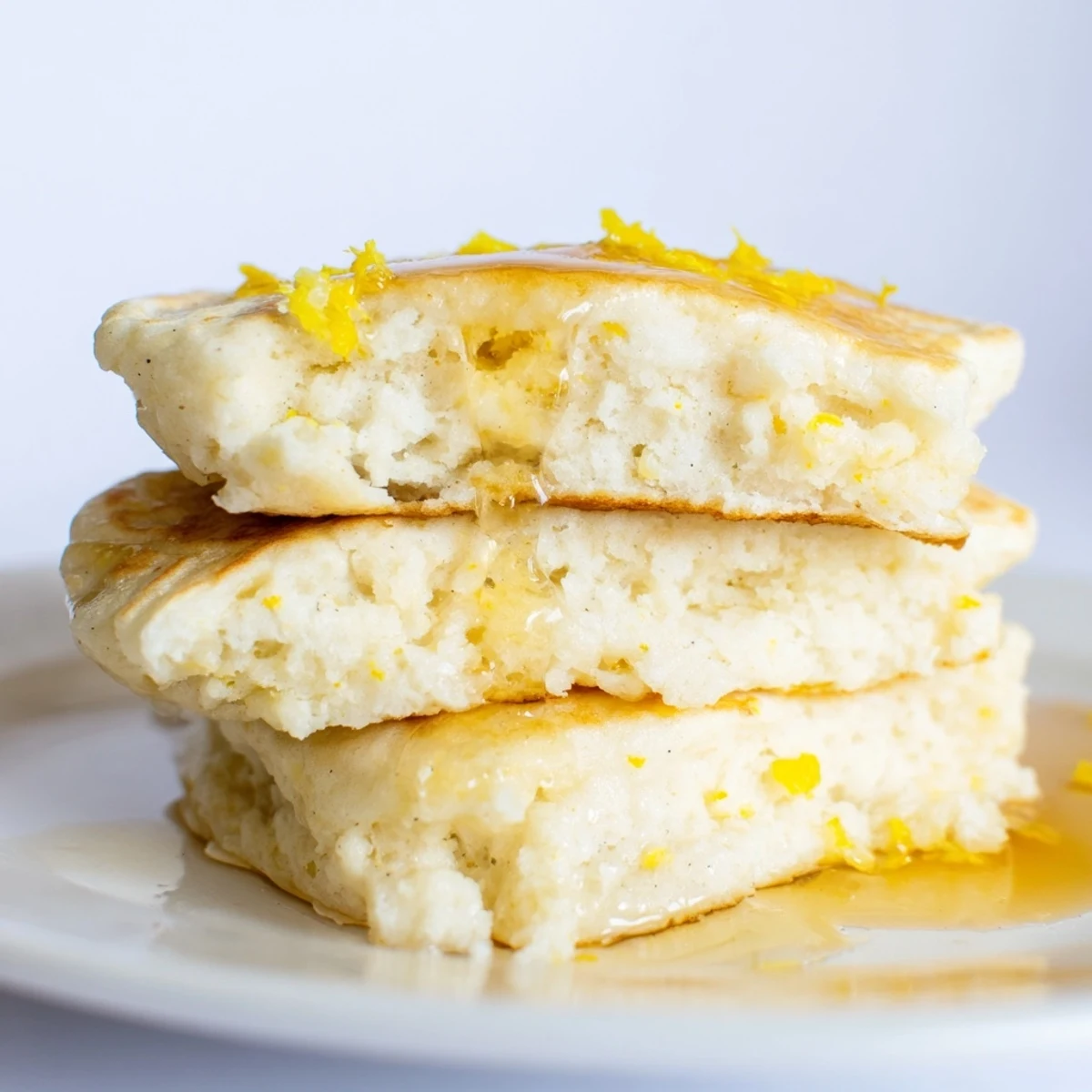 A stack of warm Lemon Ricotta Pancakes, drizzled with maple syrup and garnished with lemon zest.