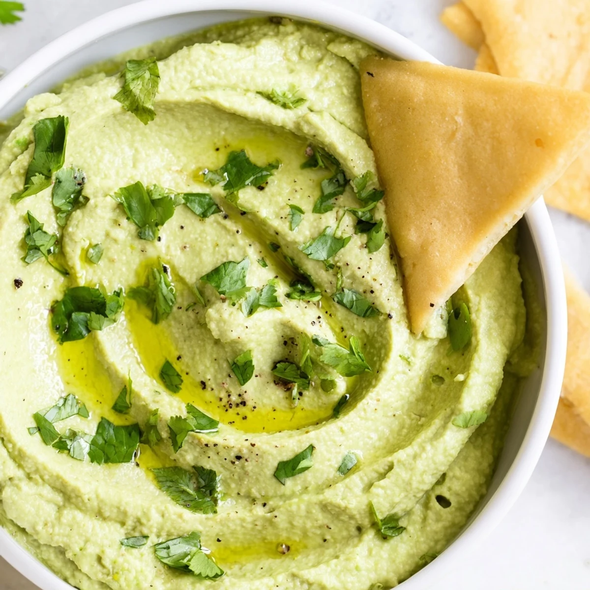 Creamy green avocado hummus dip topped with olive oil, served with golden crispy baked pita chips for dipping.