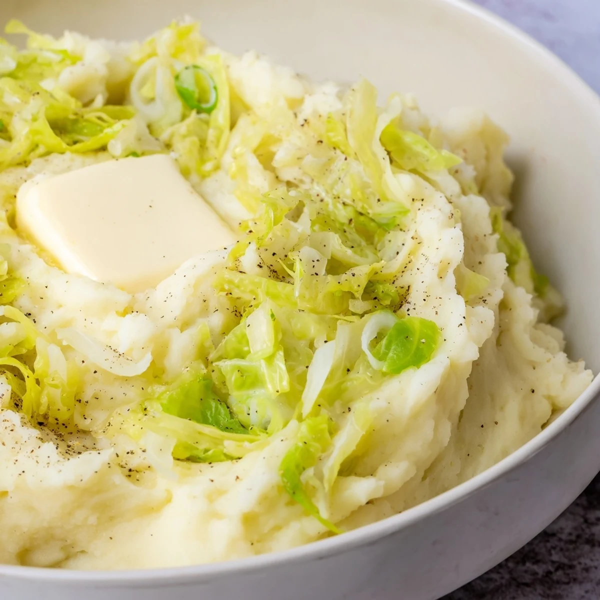 A warm bowl of Irish Colcannon Mashed Potatoes featuring fluffy spuds, tender green cabbage, and scallions, topped with a melting pat of golden butter.