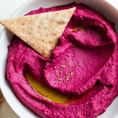 A bowl of creamy Roasted Beet Hummus garnished with sesame seeds, served alongside crunchy baked pita triangles.