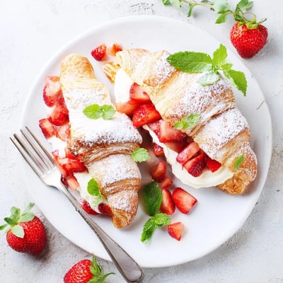 A close-up of Strawberry Cream Croissant, dusted with powdered sugar and garnished with fresh mint on a rustic board.